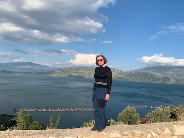 A woman in dark blue standing in front of a lake in Albania, with blue sky in the background.