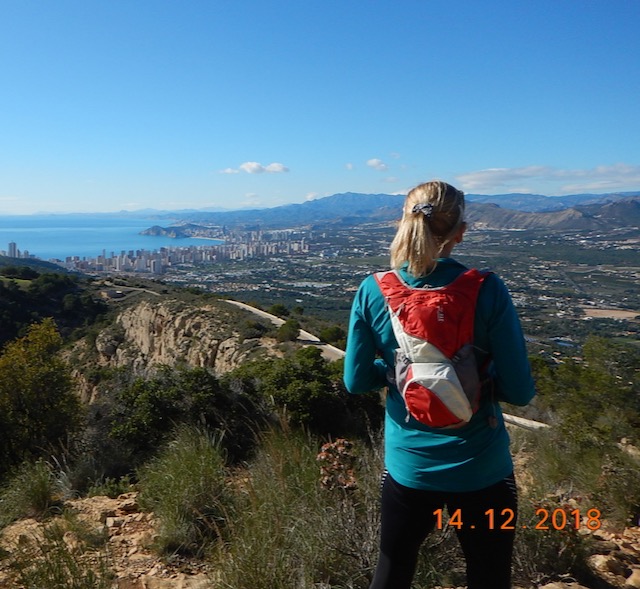A woman standing on a hipptop with a small red back pack, looking out over a town towards the blue sea with a blue sky in the background.