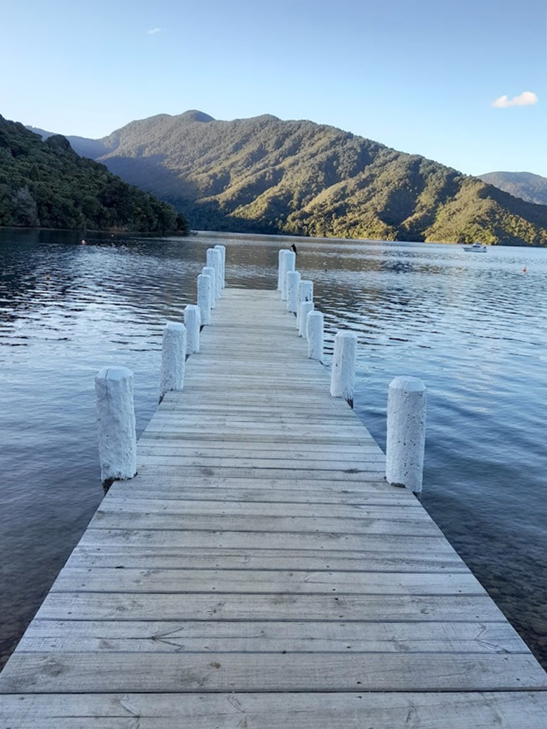 Looking down a wooden jetty with white posts, with a lake and a green hill in the distance.