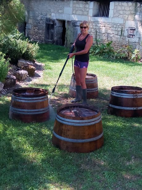 A woman in a black top and sunglasses using a high-pressure washer to clean old wine barrels.