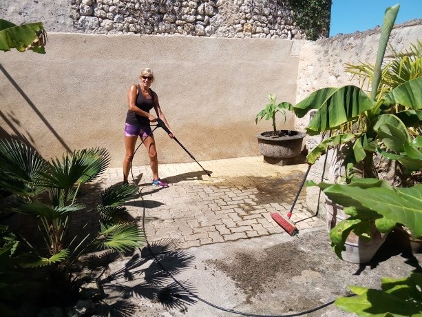 A woman in a sunny courtyard cleaning the floor paving with a high pressure washer.
