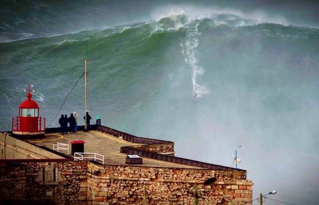 Man surfing down huge wave in front of obervers at Nazaré, Portugal